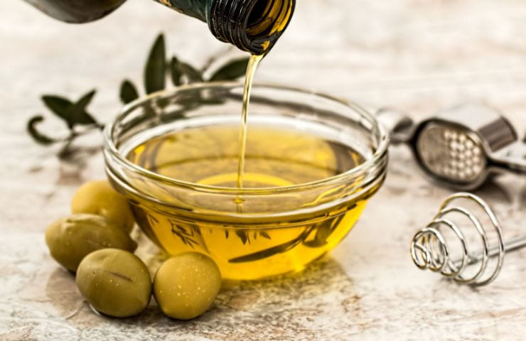 bottiglia di olio che ne versa un goccio in un contenitore già colmo di olio con vicino olive